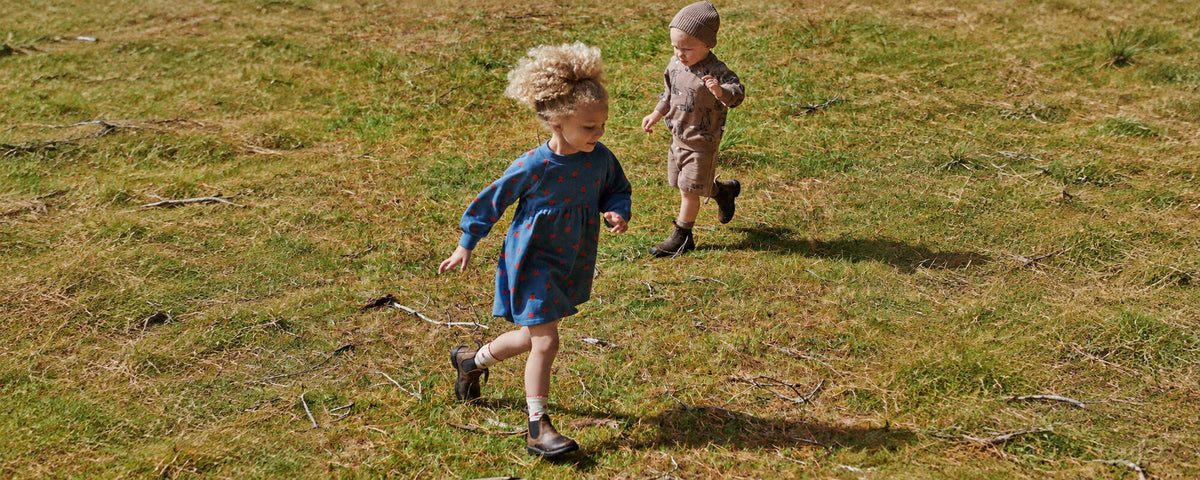 Two young children running across a grassy field in organic cotton clothing, one in a blue dress with red dots and one in a printed sweatshirt, shorts and beanie